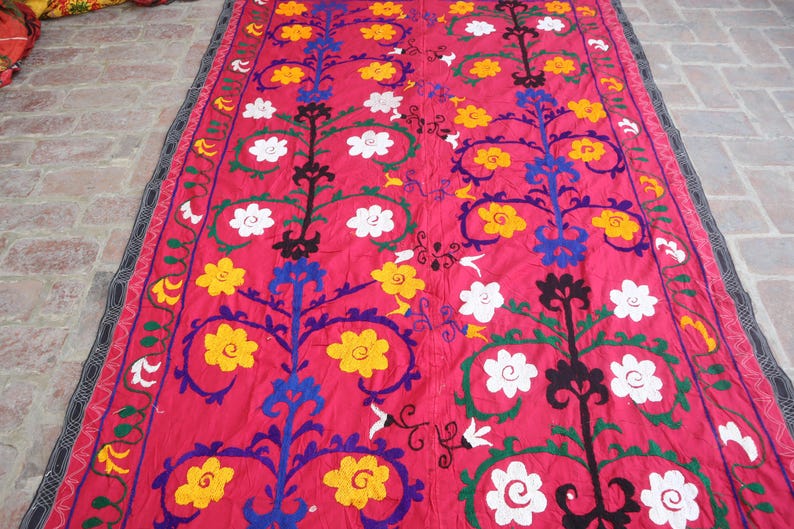K&ouml;nnte beinhalten: Ein rechteckiger Textil mit pinkfarbenem Hintergrund. Das Design zeigt stilisierte Blumenmuster in Gelb, Wei&szlig;, Blau und Gr&uuml;n. Die R&auml;nder sind mit einem schwarz-grauen Muster eingefasst. Dieses dekorative Textil eignet sich f&uuml;r die Inneneinrichtung.