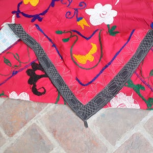K&ouml;nnte beinhalten: Ein leuchtend rotes Textil mit aufwendigen Stickereien in Wei&szlig;, Gelb, Blau und Schwarz. Das Design zeigt florale und geometrische Muster mit einem detaillierten Rand. Der Stoff ist gefaltet und zeigt die detaillierte Handwerkskunst.