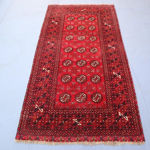May include: A rectangular red rug with an intricate geometric pattern. The rug features a central design of repeating circular motifs in brown and cream, framed by detailed borders. The rug's color palette includes shades of red, brown, and cream.