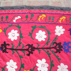 K&ouml;nnte beinhalten: Nahaufnahme eines leuchtend rosa Textils mit aufwendiger Stickerei. Das Design zeigt wei&szlig;e, gelbe und schwarze Blumenmuster sowie gr&uuml;ne und blaue Akzente. Oben befindet sich eine dekorative Bord&uuml;re mit einem schwarz-wei&szlig;en Muster.