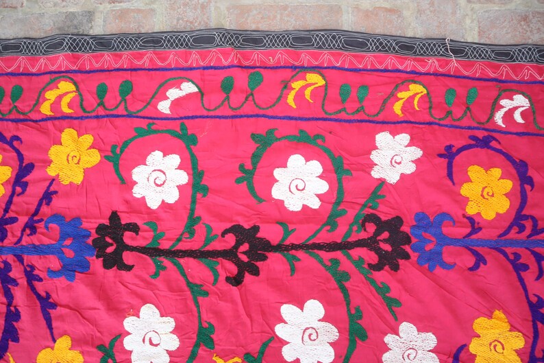 K&ouml;nnte beinhalten: Nahaufnahme eines leuchtend rosa Textils mit aufwendiger Stickerei. Das Design zeigt wei&szlig;e, gelbe und schwarze Blumenmuster sowie gr&uuml;ne und blaue Akzente. Oben befindet sich eine dekorative Bord&uuml;re mit einem schwarz-wei&szlig;en Muster.