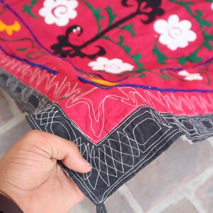 K&ouml;nnte beinhalten: Ausschnitt eines roten Textils mit wei&szlig;en Blumenstickereien und schwarzen und gr&uuml;nen Akzenten. Der Rand ist schwarz mit wei&szlig;en geometrischen N&auml;hten. Das Textil wird von einer Hand gehalten.