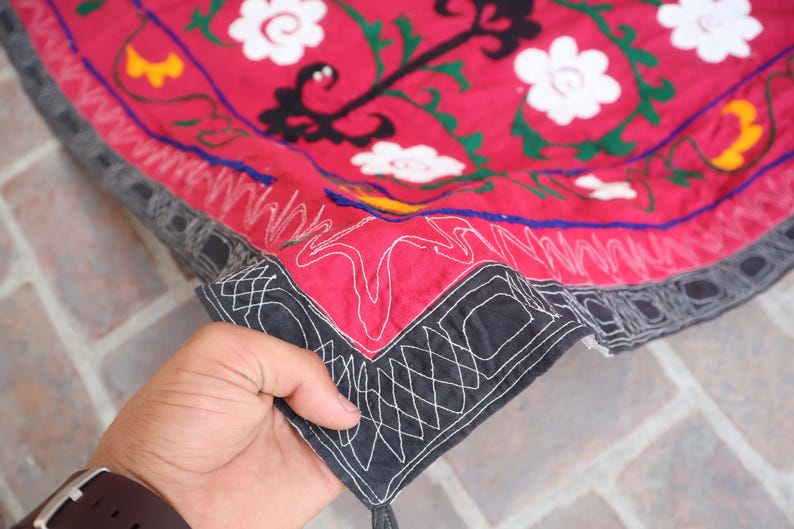 K&ouml;nnte beinhalten: Ausschnitt eines roten Textils mit wei&szlig;en Blumenstickereien und schwarzen und gr&uuml;nen Akzenten. Der Rand ist schwarz mit wei&szlig;en geometrischen N&auml;hten. Das Textil wird von einer Hand gehalten.