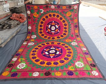 May include: A large, rectangular textile with a deep rose background. The textile features two large, circular floral designs in the centre, surrounded by smaller floral patterns in white, yellow, and green. The edges are bordered with a dark grey trim.