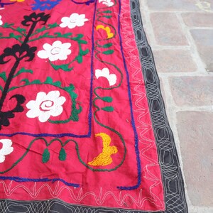 K&ouml;nnte beinhalten: Ein leuchtend rotes Textil mit gestickten Blumen- und Baumdesigns. Die Stickerei verwendet wei&szlig;e, gelbe, blaue, gr&uuml;ne und schwarze F&auml;den. Ein schwarzer Rand mit wei&szlig;en N&auml;hten umrahmt den Rand des Textils.