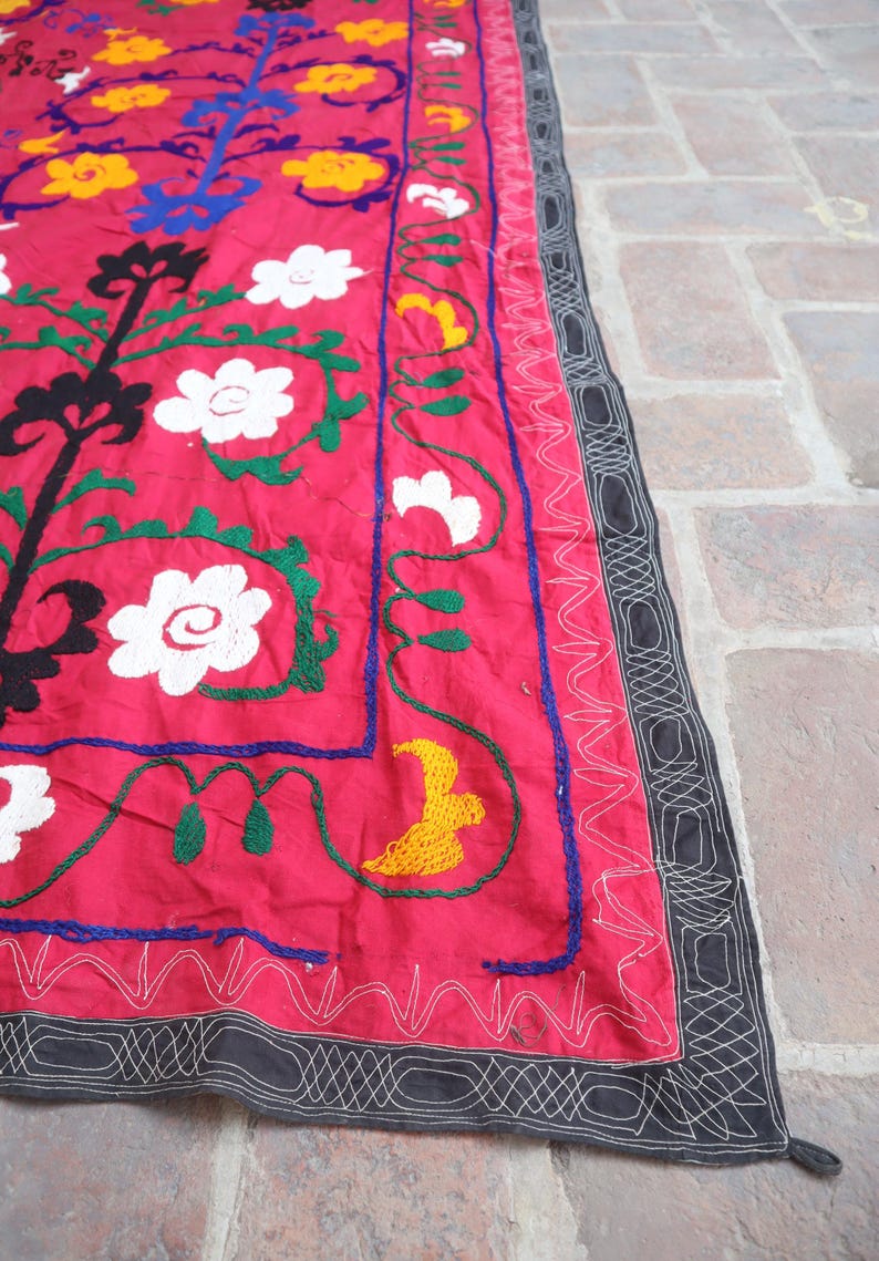 K&ouml;nnte beinhalten: Ein leuchtend rotes Textil mit gestickten Blumen- und Baumdesigns. Die Stickerei verwendet wei&szlig;e, gelbe, blaue, gr&uuml;ne und schwarze F&auml;den. Ein schwarzer Rand mit wei&szlig;en N&auml;hten umrahmt den Rand des Textils.