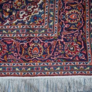 Puede incluir: Una alfombra rectangular con un campo azul marino profundo con intrincados patrones florales y de enredaderas en tonos rojo, crema, azul claro y dorado. La alfombra tiene un borde decorativo ancho y flecos de borlas blancas en el borde.