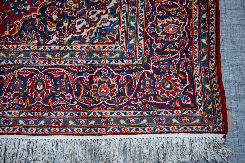 Puede incluir: Una alfombra rectangular con un campo azul marino profundo con intrincados patrones florales y de enredaderas en tonos rojo, crema, azul claro y dorado. La alfombra tiene un borde decorativo ancho y flecos de borlas blancas en el borde.