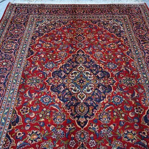 Puede incluir: Una alfombra rectangular que presenta un campo rojo vibrante con un medall&oacute;n central azul oscuro. El dise&ntilde;o incluye intrincados motivos florales y de enredaderas en tonos azul, crema y dorado, rodeado por un borde ancho y detallado.