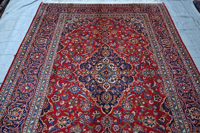 Puede incluir: Una alfombra rectangular que presenta un campo rojo vibrante con un medall&oacute;n central azul oscuro. El dise&ntilde;o incluye intrincados motivos florales y de enredaderas en tonos azul, crema y dorado, rodeado por un borde ancho y detallado.