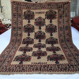 May include: A beige rug with a repeating tree design in shades of brown, red, and blue. The rug has a detailed border and fringe along the edges. Other rugs are visible in the background.