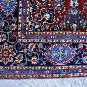 K&ouml;nnte beinhalten: Ein rechteckiger Teppich mit einem tiefroten Mittelfeld und einem breiten marineblauen Rand. Das Design umfasst komplexe florale Motive, stilisierte Ranken und geometrische Formen in hellblauen, cremefarbenen und beigen T&ouml;nen, abgeschlossen mit wei&szlig;en Fransen.