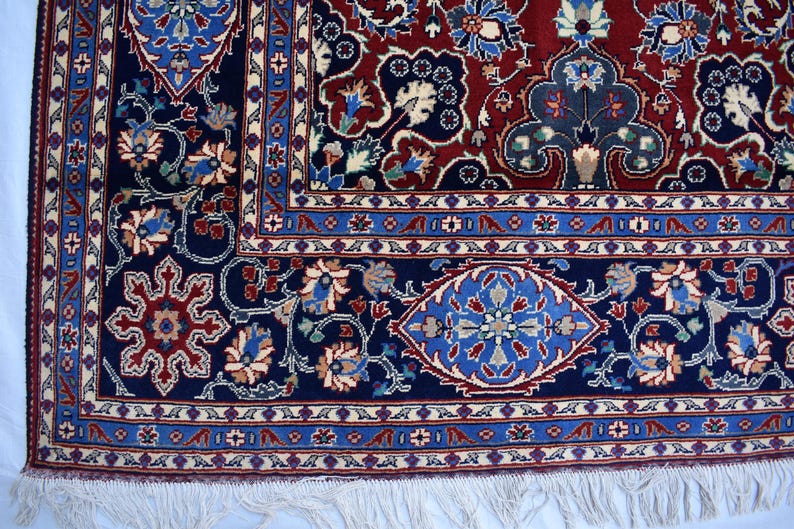 K&ouml;nnte beinhalten: Ein rechteckiger Teppich mit einem tiefroten Mittelfeld und einem breiten marineblauen Rand. Das Design umfasst komplexe florale Motive, stilisierte Ranken und geometrische Formen in hellblauen, cremefarbenen und beigen T&ouml;nen, abgeschlossen mit wei&szlig;en Fransen.