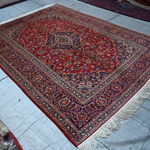 Puede incluir: Una alfombra rectangular que presenta un campo central de color rojo intenso con un medall&oacute;n azul oscuro y patrones florales intrincados en tonos crema, azul claro y dorado. La alfombra tiene un borde ancho azul oscuro con flecos blancos.