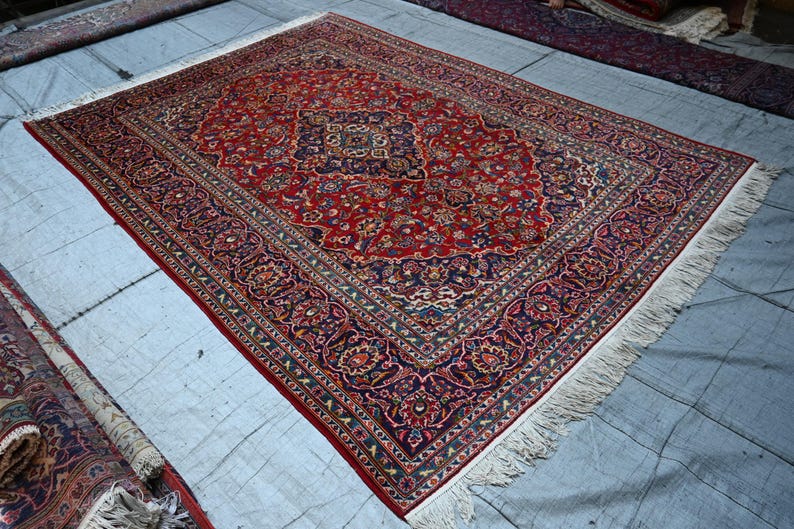 Puede incluir: Una alfombra rectangular que presenta un campo central de color rojo intenso con un medall&oacute;n azul oscuro y patrones florales intrincados en tonos crema, azul claro y dorado. La alfombra tiene un borde ancho azul oscuro con flecos blancos.