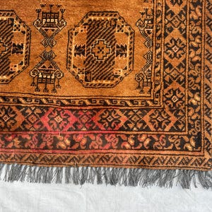 May include: A hand-knotted rug featuring a warm orange field with repeating dark brown octagonal geometric motifs and stylized floral elements. The border displays intricate dark brown and red patterns, finished with a grey fringed edge.