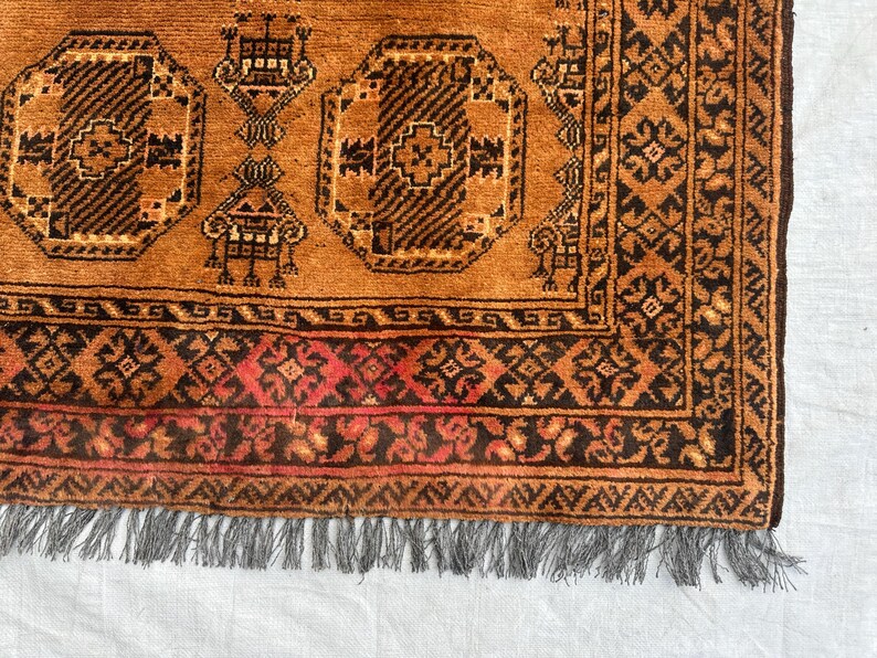 May include: A hand-knotted rug featuring a warm orange field with repeating dark brown octagonal geometric motifs and stylized floral elements. The border displays intricate dark brown and red patterns, finished with a grey fringed edge.