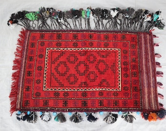 May include: A red rug with a geometric pattern and black tassels. The rug has a central square with a smaller square inside. The rug is surrounded by a border of smaller squares.
