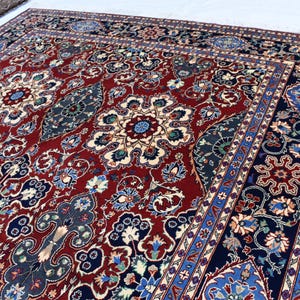 K&ouml;nnte beinhalten: Ein gro&szlig;er rechteckiger Teppich mit einem tiefroten Mittelfeld, das mit komplizierten Blumenmedaillons und geschwungenen Rankenmotiven in Marineblau, Creme und Hellblau verziert ist, umrahmt von einer breiten dunkelblauen Bord&uuml;re mit Blumenmustern.