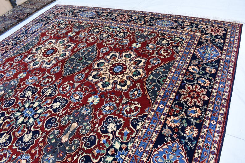 K&ouml;nnte beinhalten: Ein gro&szlig;er rechteckiger Teppich mit einem tiefroten Mittelfeld, das mit komplizierten Blumenmedaillons und geschwungenen Rankenmotiven in Marineblau, Creme und Hellblau verziert ist, umrahmt von einer breiten dunkelblauen Bord&uuml;re mit Blumenmustern.