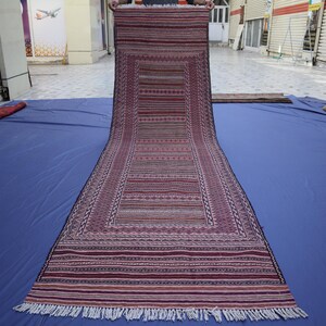 May include: A long, rectangular runner rug featuring a deep maroon and purple color palette with intricate horizontal striped patterns and geometric borders. The rug has a flat-woven texture and is finished with short white fringe at the ends.