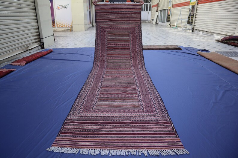 May include: A long, rectangular runner rug featuring a deep maroon and purple color palette with intricate horizontal striped patterns and geometric borders. The rug has a flat-woven texture and is finished with short white fringe at the ends.