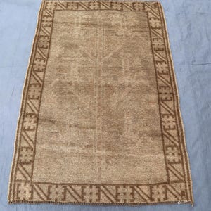 May include: Rectangular brown rug with a geometric border and central design. The rug has a faded appearance, with a lighter brown centre and darker brown border. The rug is placed on a blue surface.