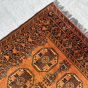 May include: A rectangular area rug featuring a burnt orange background with repeating dark brown geometric octagonal motifs and intricate border patterns. The rug is finished with a short, dark gray fringe along the edge of the woven fabric.