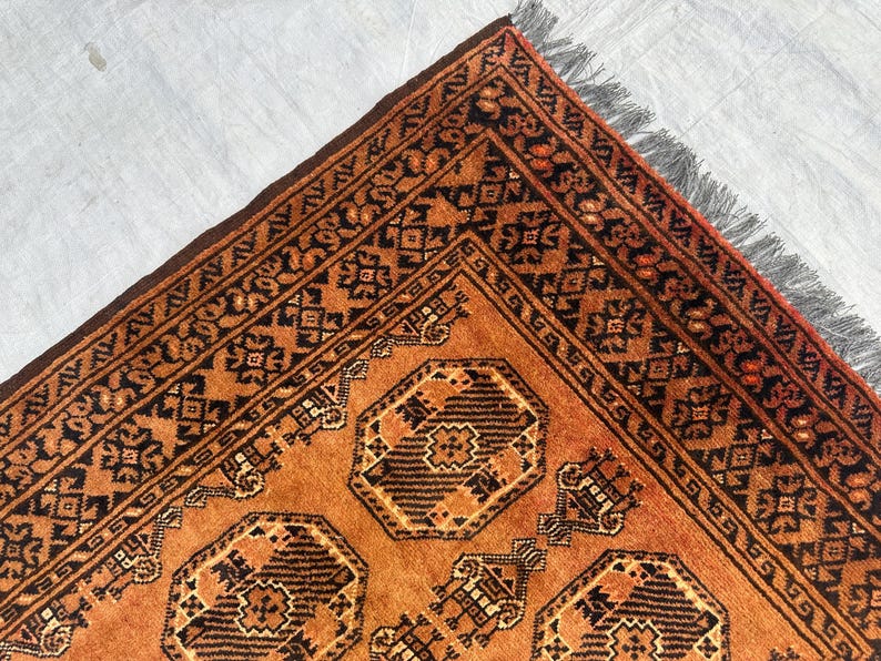 May include: A rectangular area rug featuring a burnt orange background with repeating dark brown geometric octagonal motifs and intricate border patterns. The rug is finished with a short, dark gray fringe along the edge of the woven fabric.