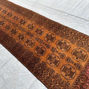 May include: A rectangular runner rug featuring a vibrant burnt orange background with repeating dark brown octagonal geometric motifs. The rug is bordered by intricate dark brown and black patterns and includes short fringe at the ends.