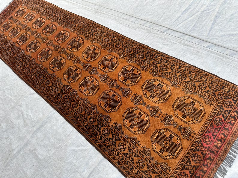 May include: A rectangular runner rug featuring a vibrant burnt orange background with repeating dark brown octagonal geometric motifs. The rug is bordered by intricate dark brown and black patterns and includes short fringe at the ends.