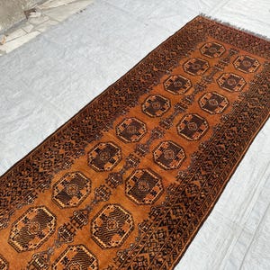 May include: A long rectangular runner rug featuring a deep orange background with repeating dark brown octagonal geometric motifs. The rug is framed by intricate dark brown borders with detailed patterns and has short fringe at the ends.