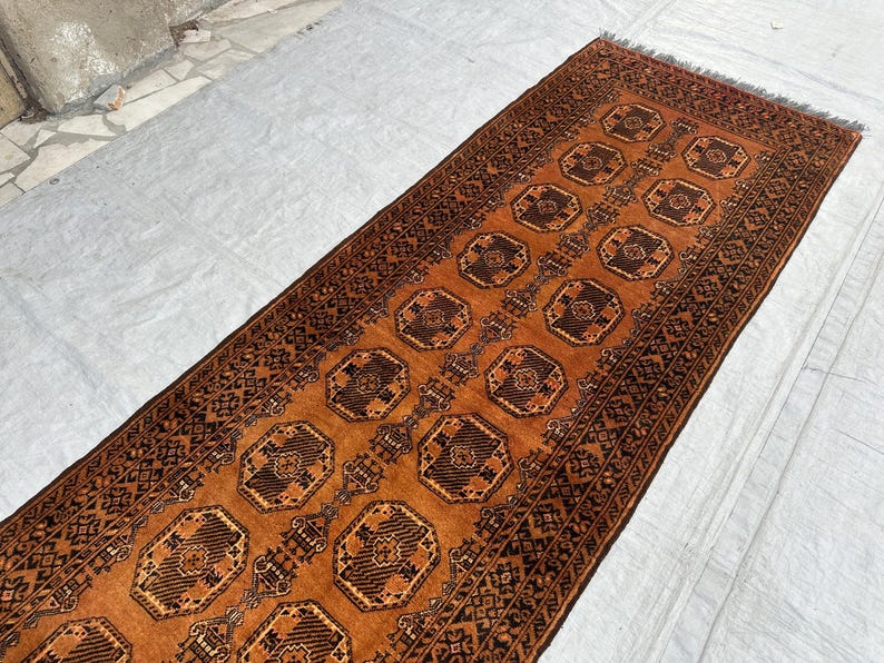May include: A long rectangular runner rug featuring a deep orange background with repeating dark brown octagonal geometric motifs. The rug is framed by intricate dark brown borders with detailed patterns and has short fringe at the ends.