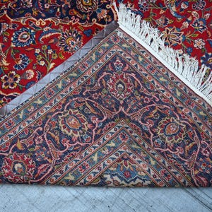 Puede incluir: Una alfombra rectangular con un complejo patr&oacute;n floral en tonos rojo, azul marino y crema. La alfombra est&aacute; doblada para mostrar un reverso azul oscuro con motivos bot&aacute;nicos detallados y un fleco blanco a lo largo del borde.