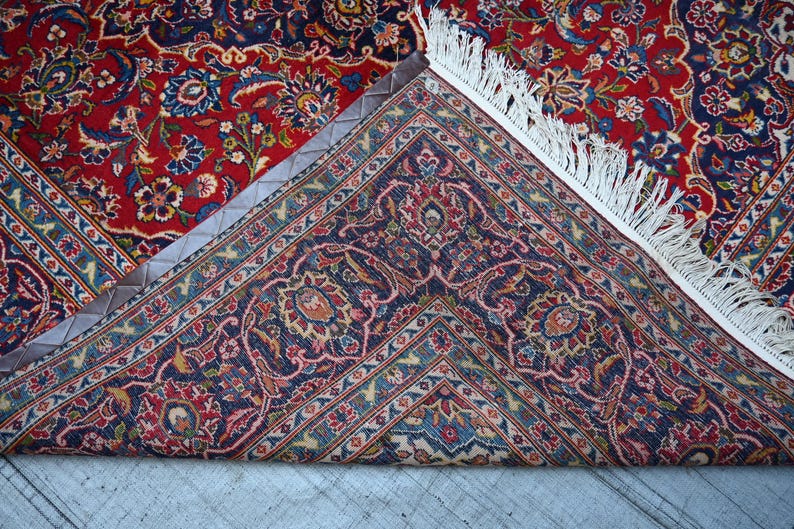 Puede incluir: Una alfombra rectangular con un complejo patr&oacute;n floral en tonos rojo, azul marino y crema. La alfombra est&aacute; doblada para mostrar un reverso azul oscuro con motivos bot&aacute;nicos detallados y un fleco blanco a lo largo del borde.