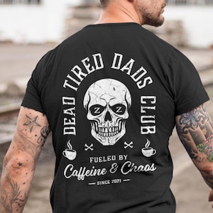 May include: Black t-shirt with a white skull graphic and the text "Dead Tired Dads Club" with a cross on either side of the skull. Below the skull is the text "Fueled by Caffeine & Chaos" with the text "Since 2021" underneath.