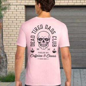May include: A light pink short-sleeve t-shirt featuring a black graphic on the back. The design includes a skull with Zs in the eye sockets, the text DEAD TIRED DADS CLUB, FUELED BY Caffeine & Chaos, and the text SINCE 2024 with coffee cups.