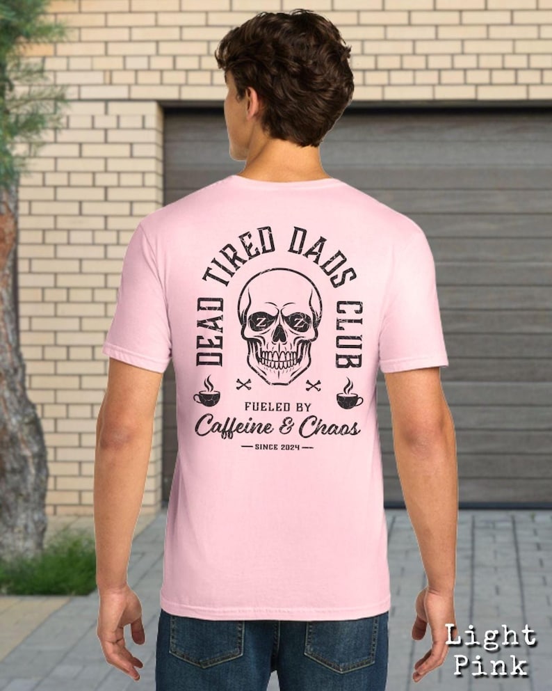 May include: A light pink short-sleeve t-shirt featuring a black graphic on the back. The design includes a skull with Zs in the eye sockets, the text DEAD TIRED DADS CLUB, FUELED BY Caffeine & Chaos, and the text SINCE 2024 with coffee cups.