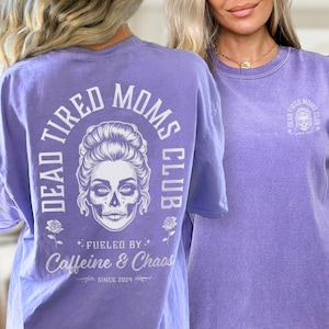 Peut inclure: T-shirt violet avec l'inscription "DEAD TIRED MOMS CLUB" et un motif de cr&acirc;ne. Le t-shirt comporte &eacute;galement les phrases "FUELED BY Caffeine & Chaos" et "SINCE 2024".