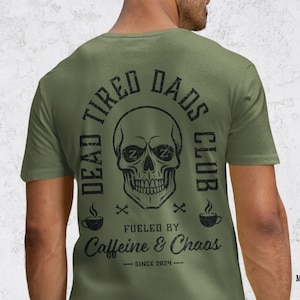May include: A military green short-sleeve t-shirt featuring a black graphic on the back. The design includes a skull with Zs in the eye sockets, surrounded by the text DEAD TIRED DADS CLUB, FUELED BY CAFFEINE & CHAOS, and SINCE 2024.