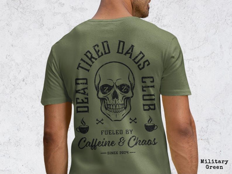 May include: A military green short-sleeve t-shirt featuring a black graphic on the back. The design includes a skull with Zs in the eye sockets, surrounded by the text DEAD TIRED DADS CLUB, FUELED BY CAFFEINE & CHAOS, and SINCE 2024.