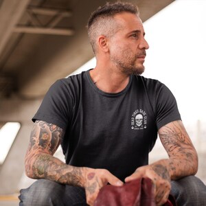 May include: A charcoal gray short-sleeve t-shirt featuring a white circular graphic on the left chest. The graphic depicts a skull with the text "DEAD TIRED DADS CLUB" arched above and "SINCE 2021" printed below the skull design.