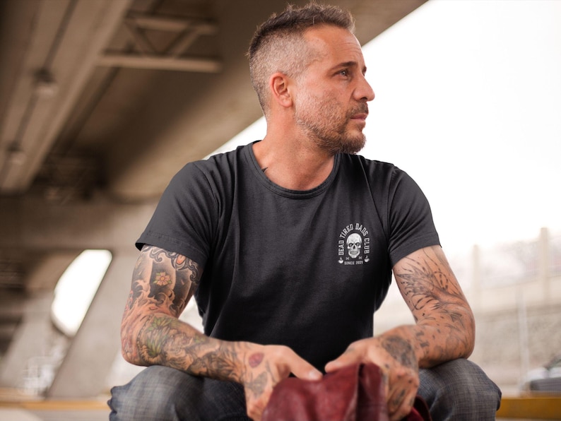 May include: A charcoal gray short-sleeve t-shirt featuring a white circular graphic on the left chest. The graphic depicts a skull with the text "DEAD TIRED DADS CLUB" arched above and "SINCE 2021" printed below the skull design.