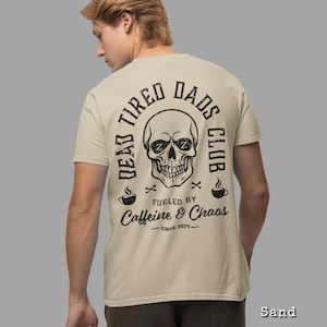 May include: A sand-colored t-shirt featuring a black graphic on the back. The design includes a skull with Zs in the eye sockets, surrounded by the text DEAD TIRED DADS CLUB, FUELED BY CAFFEINE & CHAOS, and SINCE 2024, with two coffee cups.