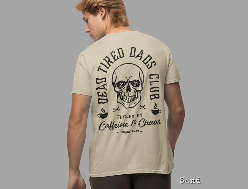 May include: A sand-colored t-shirt featuring a black graphic on the back. The design includes a skull with Zs in the eye sockets, surrounded by the text DEAD TIRED DADS CLUB, FUELED BY CAFFEINE & CHAOS, and SINCE 2024, with two coffee cups.