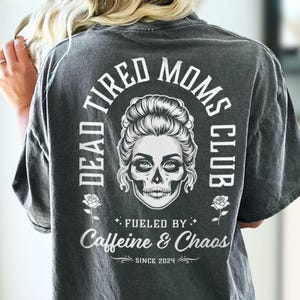 Peut inclure: T-shirt gris fonc&eacute; avec un motif au dos. Le design pr&eacute;sente un cr&acirc;ne avec une coiffure f&eacute;minine, entour&eacute; des mots "DEAD TIRED MOMS CLUB" et "Fueled by Caffeine & Chaos Since 2024".