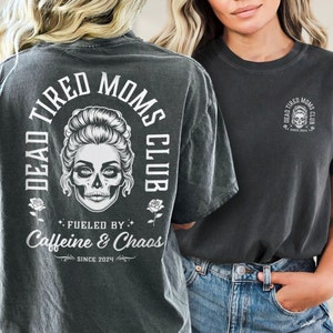 Peut inclure: T-shirt gris fonc&eacute; avec un motif de cr&acirc;ne et le texte "DEAD TIRED MOMS CLUB". Le dos du t-shirt affiche les mots "Fueled by Caffeine & Chaos" et "Since 2024". Le devant pr&eacute;sente une version plus petite du motif.