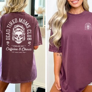 Peut inclure: T-shirt de couleur baie avec l'inscription "DEAD TIRED MOMS CLUB" et un motif de cr&acirc;ne. Le dos du t-shirt porte l'inscription "Fueled by Caffeine & Chaos Since 2024". Le devant du t-shirt pr&eacute;sente une version plus petite du motif de cr&acirc;ne.