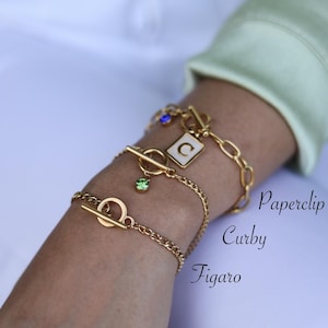 May include: Three gold chain bracelets with different link styles. The top bracelet has a charm with the letter "C" and a small blue stone. The middle bracelet has a green stone charm. The bottom bracelet has a toggle clasp. The bracelets are labeled "Paperclip", "Curby", and "Figaro".