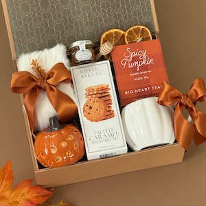 May include: A gift box containing a white faux fur item with a brown ribbon, a small jar of honey with a wooden dipper, a box of Rustic Bakery salted caramel shortbread, a box of Big Heart Tea Spicy Pumpkin tea, and a white pumpkin-shaped mug.